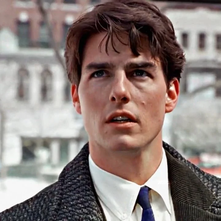 A young man with dark hair and fair skin, wearing a white shirt, dark tie, and a dark coat, looking slightly confused or surprised in an outdoor setting with blurred buildings in the background.