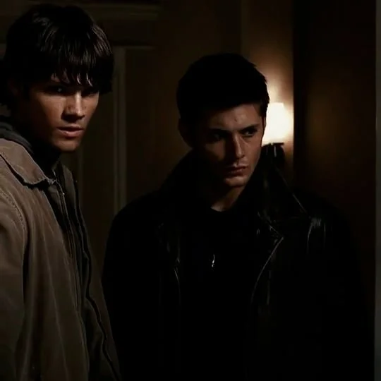 Two young men standing indoors in a dimly lit room, one wearing a beige jacket and the other in a black leather jacket.