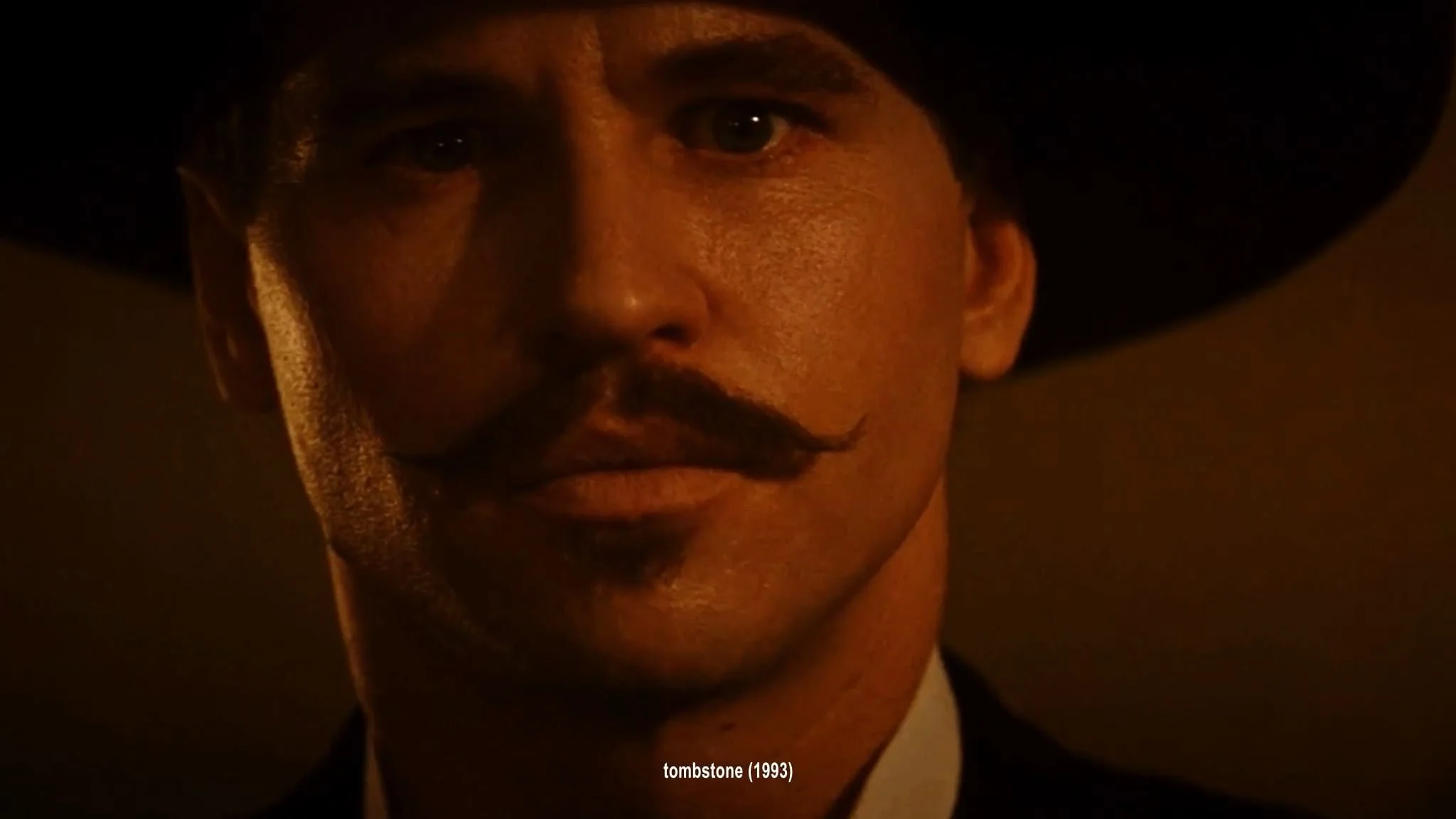 Close-up of a man with a mustache wearing a wide-brimmed hat, dark background, movie still from 'Tombstone' (1993).