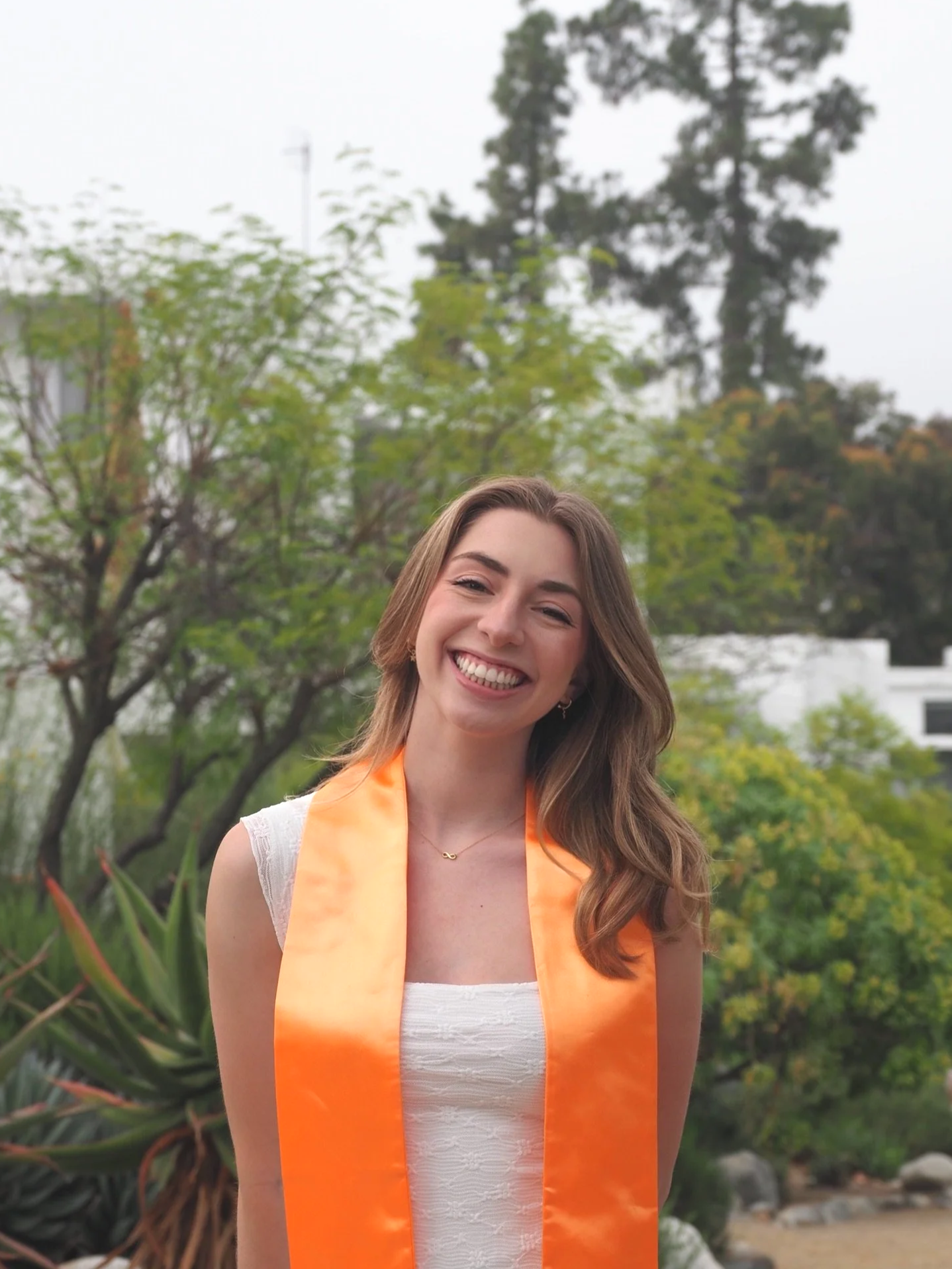 A young woman smiling and wearing an orange graduation stole outdoors, with green trees and plants in the background.