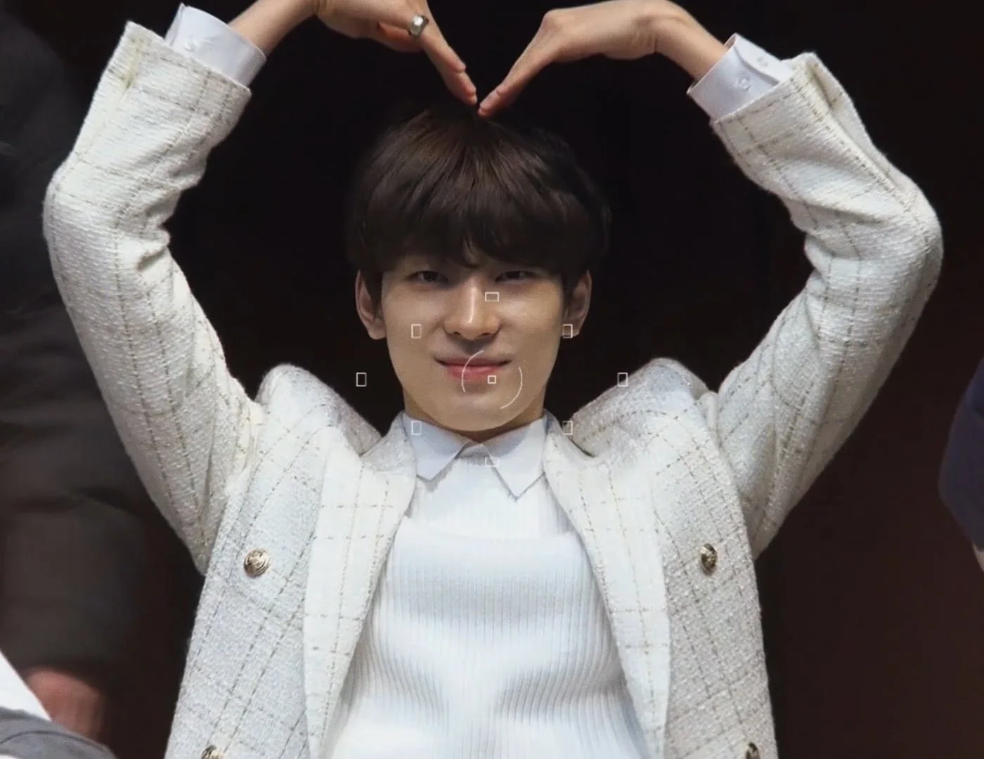 A young man lying down on a dark surface, making a heart shape with his hands above his head, smiling at the camera. He is wearing a white sweater and a textured white blazer.