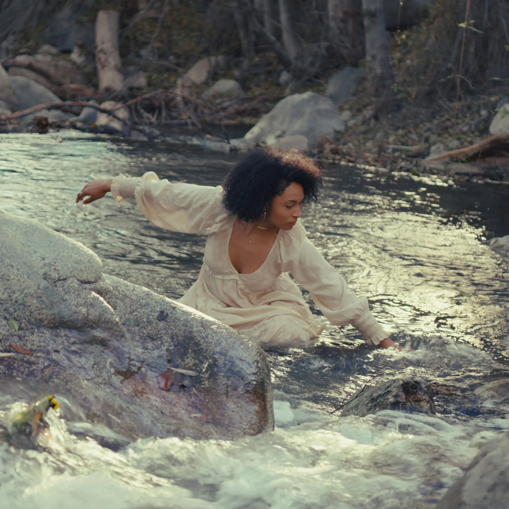 Leah Lahkiah with curly dark hair and wearing a white dress is squatting on rocks playing inside a body of water in a natural outdoor setting with trees and rocks in the background.
