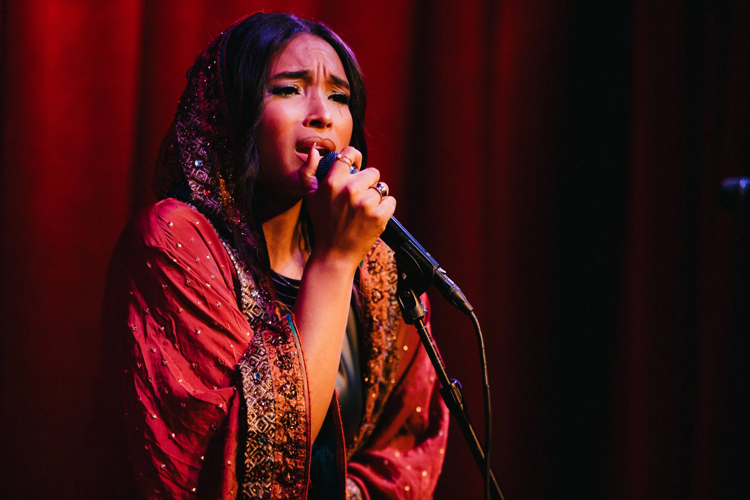 Leah Lahkiah  with long dark hair singing into microphone on iconic Los Angeles hotel cafe stage. She is wearing a red hooded and decorated cardigan and appears emotional, with a jewel tear on her face. The background is dark with red curtains.