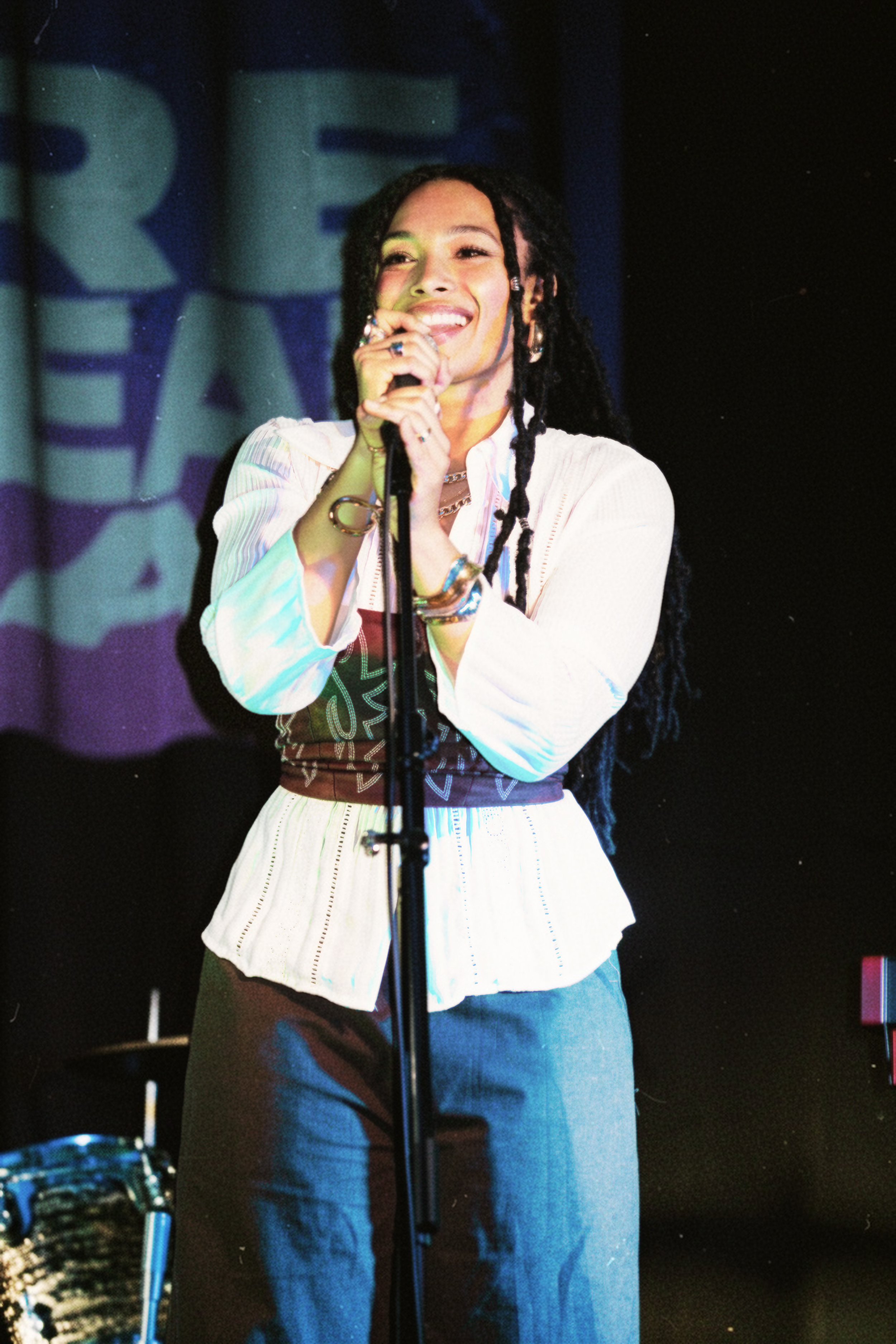 Leah Lahkiah with long faux locs smiling and singing into a microphone on Los Angeles event "The Rehearsal" stage, wearing a creamy shirt and brown pants, with a background showing large blurred "Rehearsal" letters.