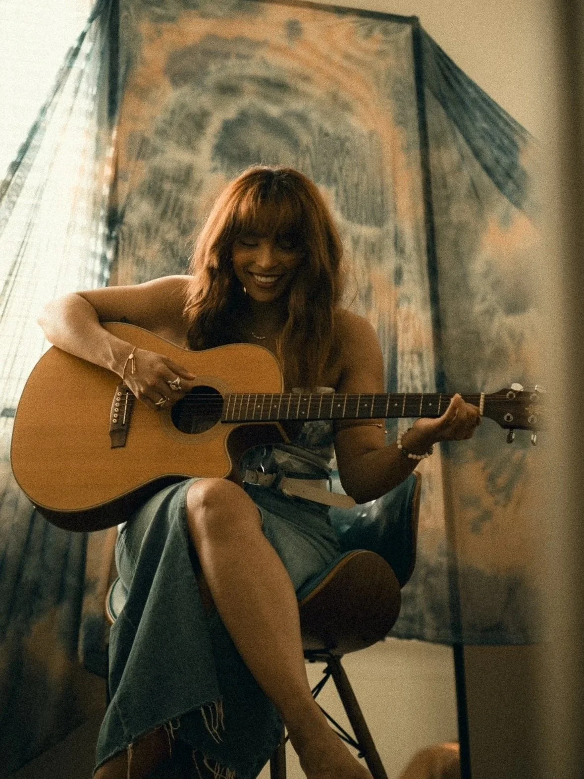 Leah Lahkiah with long auburn hair playing an acoustic guitar and smiling, sitting on a chair with fashionable denim skirt, in front of a muted abstract backdrop.
