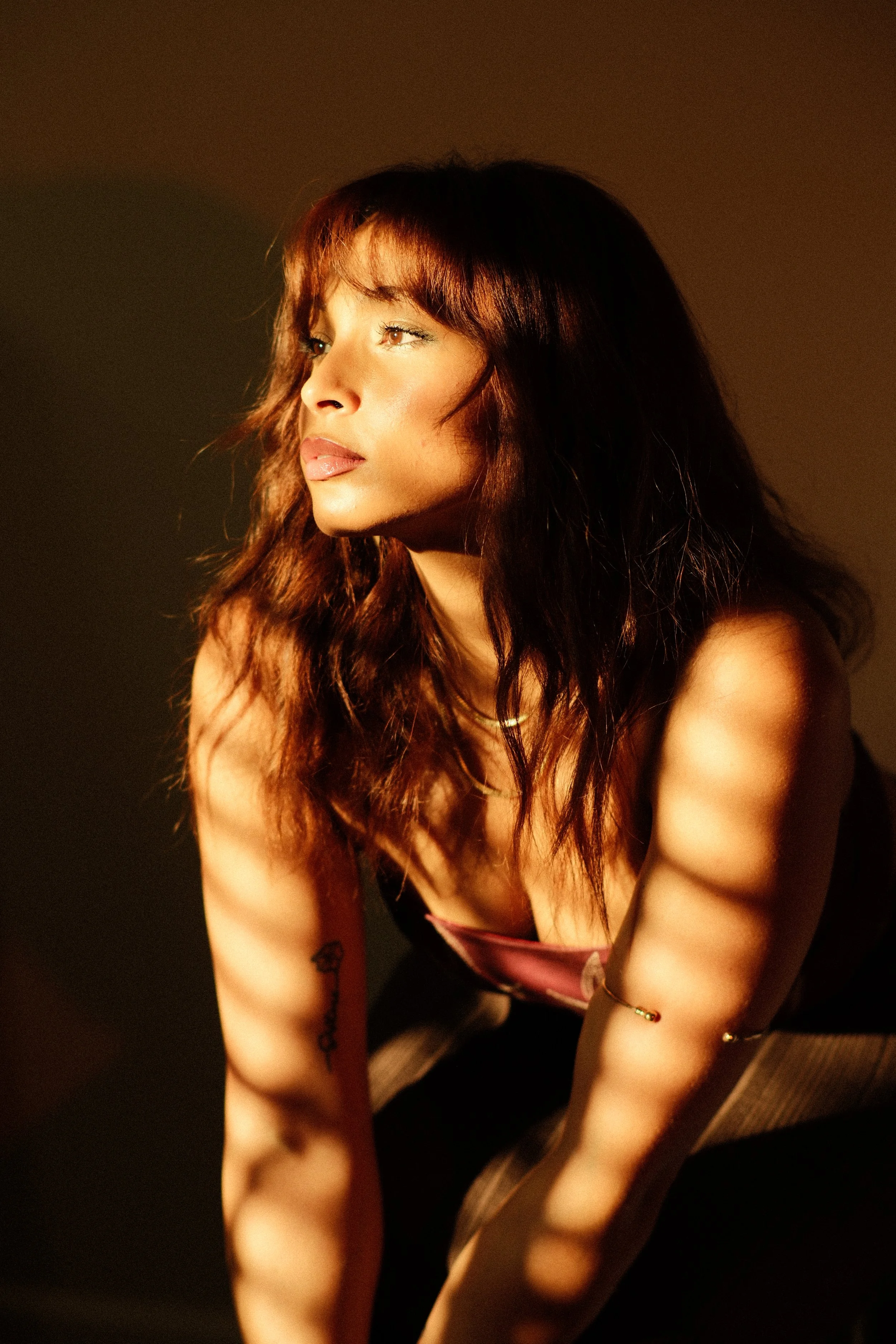Leah Lahkiah with wavy auburn hair gazing to the side out of the window , sitting on a wooden floor, illuminated by warm lighting, wearing a pink and black top, with tattoos on her right arm.