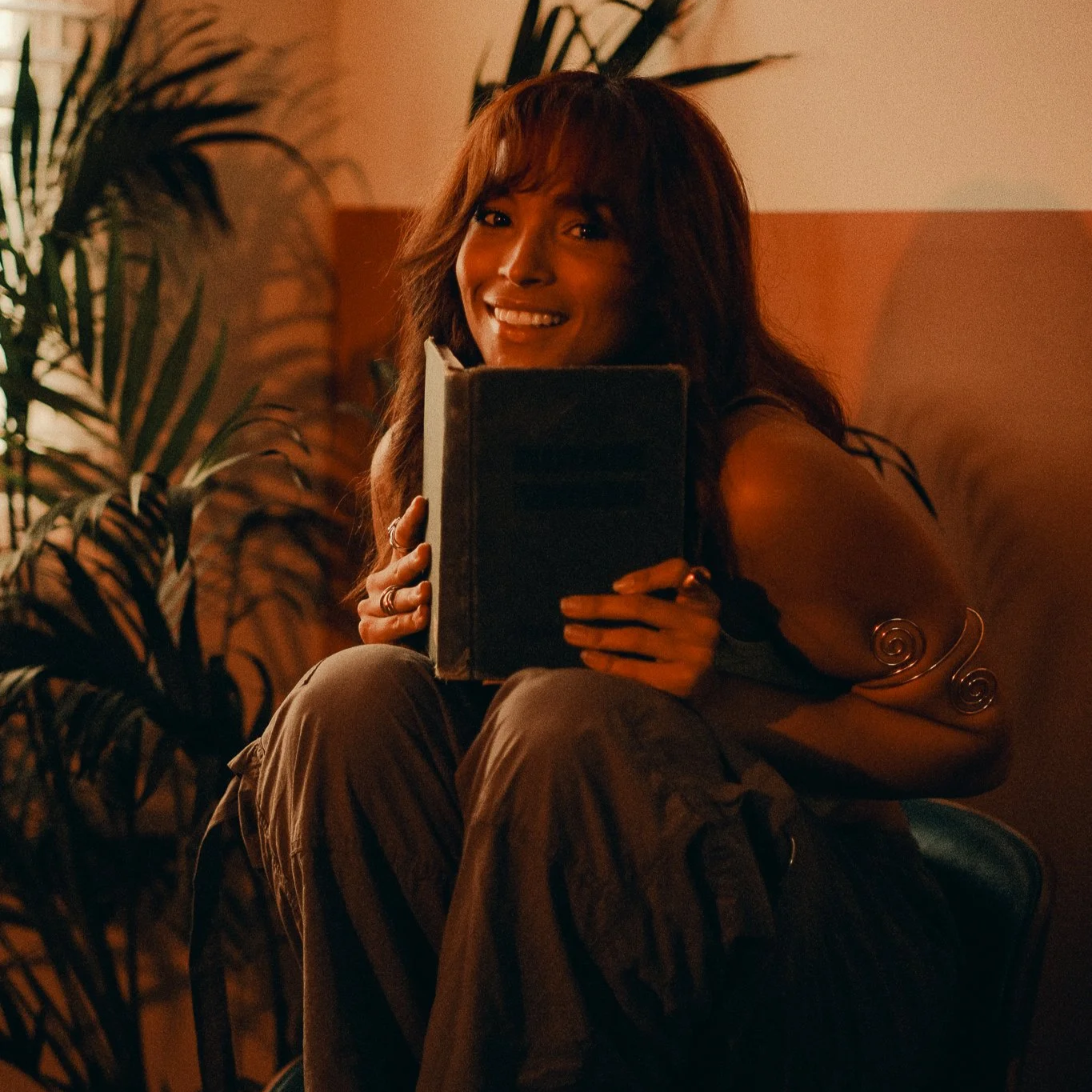 Leah Lahkiah  with reddish-brown hair smiling while holding a green hymn song book, sitting among leafy plants in a warmly lit room.