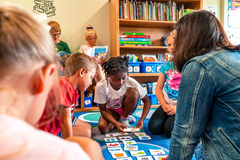 A group of young children sitting on a blue circular rug in a classroom, with one girl in the center engaging in a card matching activity while an adult assists. Other children are watching or reading books, and bookshelves with colorful books are in the background.