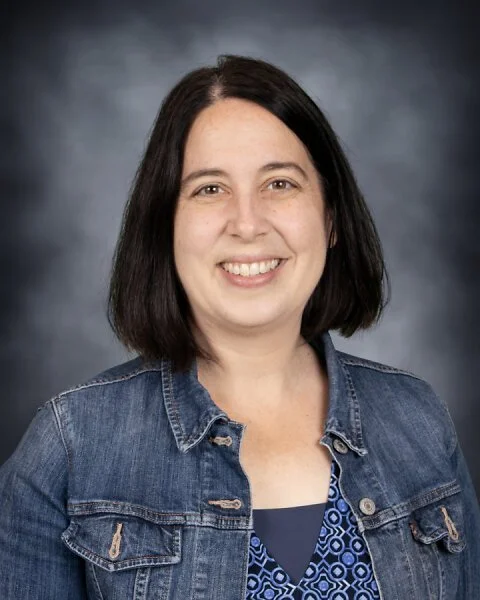 A woman with shoulder-length dark hair, smiling, wearing a denim jacket over a blue patterned top, posed against a dark gray background.