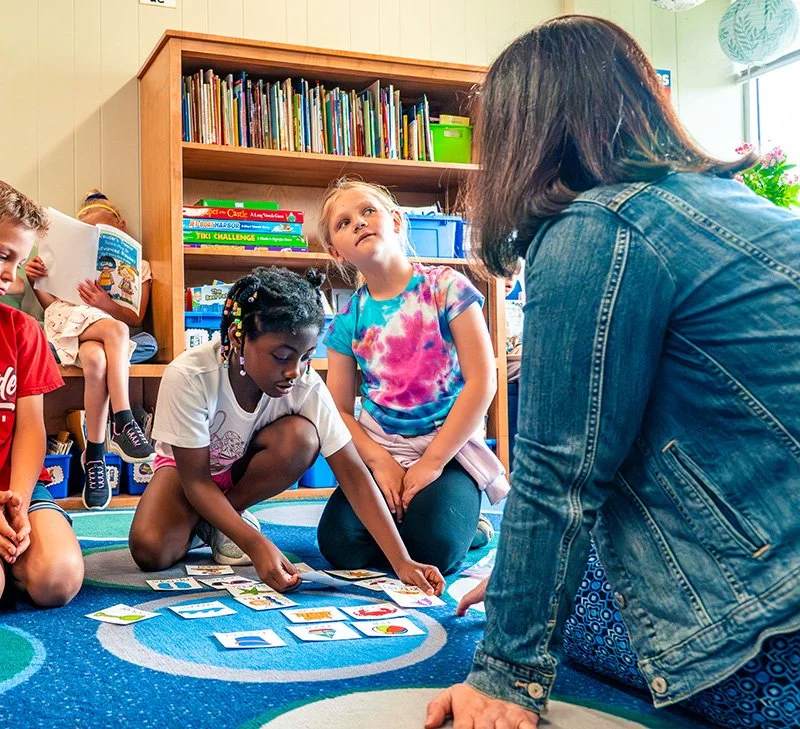 Children sitting on a colorful carpet inside a classroom, playing an educational card game with a teacher or caregiver.