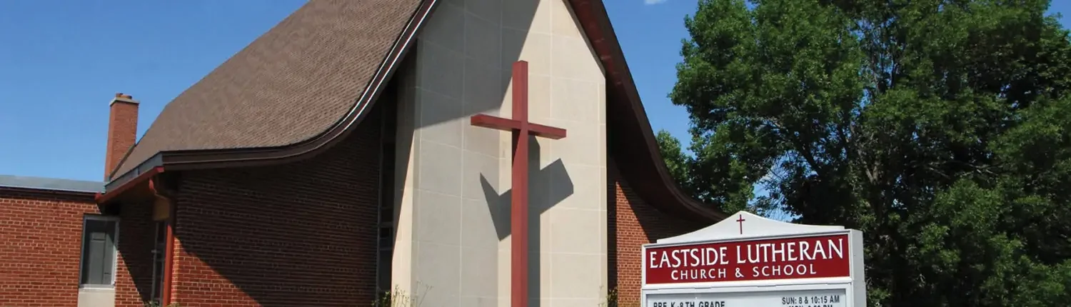 A church with a large wooden cross on the front and a white sign reading 'Eastside Lutheran Church & School' in front of green trees and a clear blue sky.