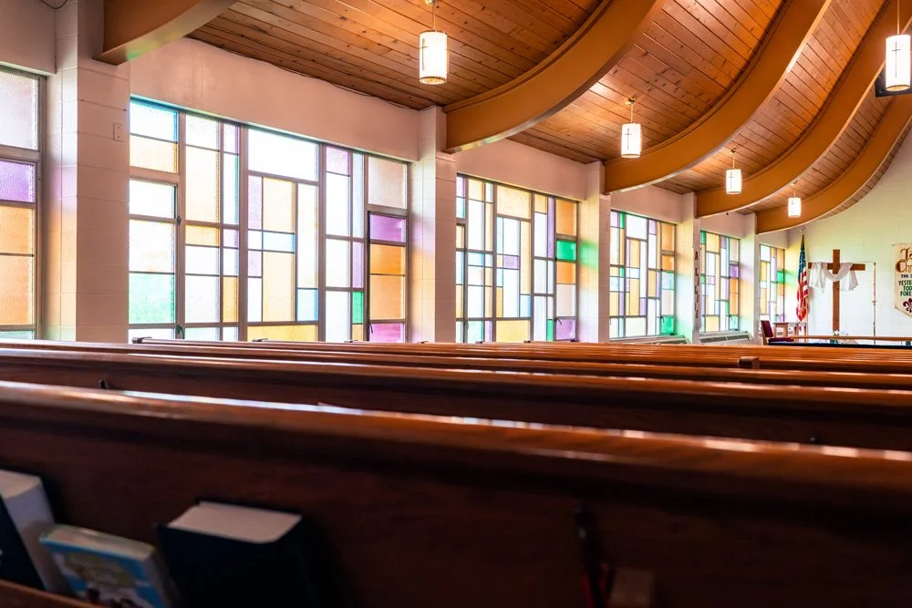 Interior of a church with wooden pews, colorful stained glass windows, hanging lights, a cross, and an American flag.