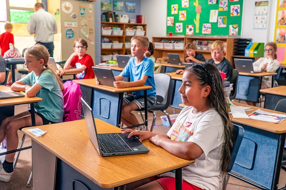 Elementary school classroom with students using laptops at their desks, colorful artwork on the walls, and a teacher at the front.