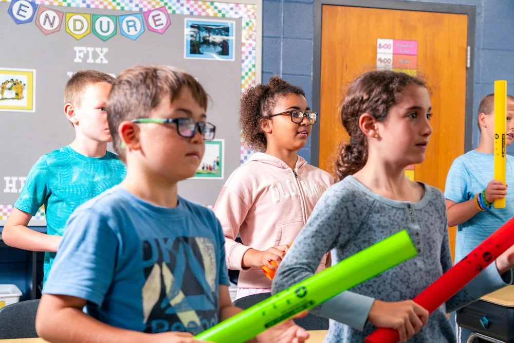 Group of children standing in a classroom holding colorful poster tubes, with a bulletin board in the background.