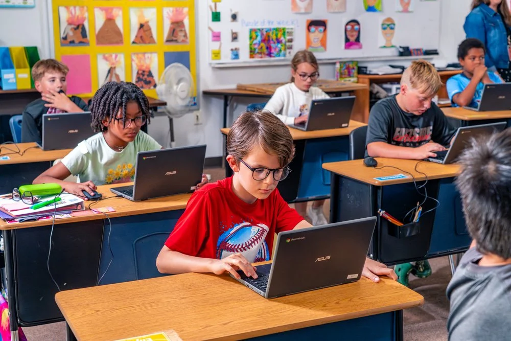 Classroom with children working on laptops at desks, decorated with colorful artwork and posters on the walls.