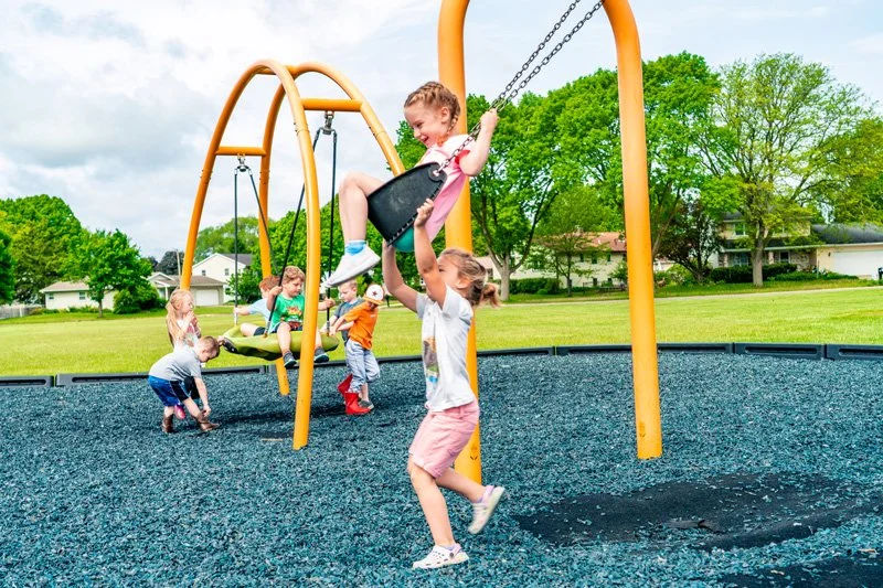 Children playing on swings at a park surrounded by green trees and residential houses on a partly cloudy day.