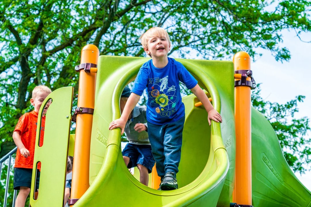 Children playing on a green slide at a playground, with trees in the background, joyful expressions on their faces.