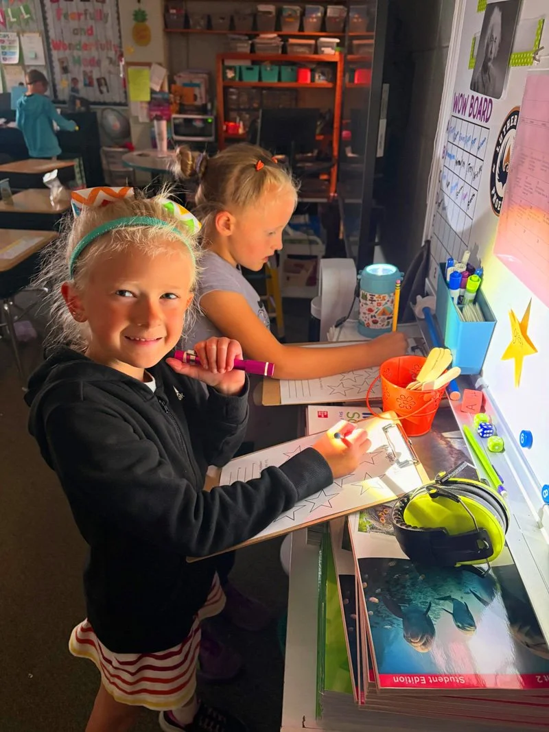 Two young girls working on school assignments at a classroom desk, with one smiling and holding a pink marker, the other focused on writing, surrounded by school supplies and educational posters.