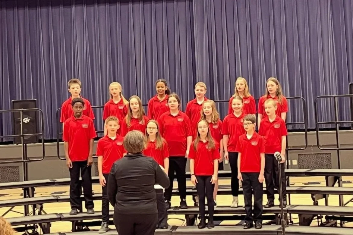 Young children in red shirts singing on stage with a conductor in front of them and a blue curtain backdrop.