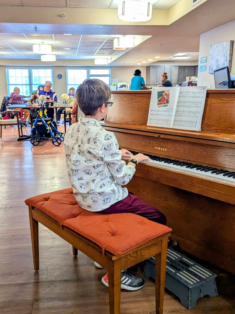 A young boy with glasses, wearing a patterned shirt and maroon pants, sitting on a padded bench, playing a Yamaha upright piano in a shared common area. Sheet music is open on the piano.