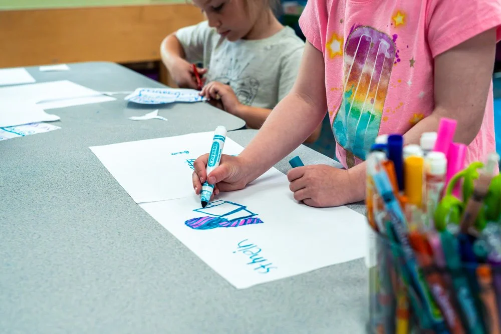 A child drawing on a large white sheet of paper with various colored markers, with a container of markers nearby, while another child in the background also draws at the table.