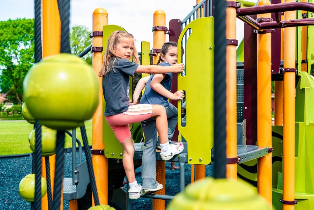 Two young girls playing on a colorful playground structure with green, orange, and purple elements, surrounded by a grassy field and trees in the background.