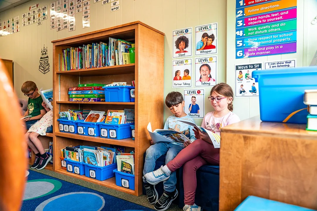 Three children sitting in a classroom reading books, with a wooden bookshelf filled with books and educational posters on the wall.