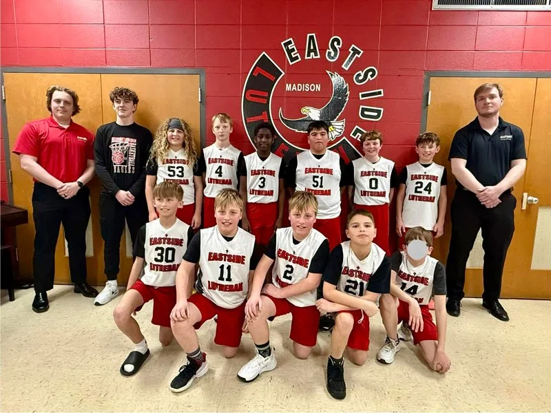 A youth basketball team in uniform, along with two coaches, posing in front of a basketball team logo on a red wall. The team is wearing white jerseys with black sleeves and red shorts.