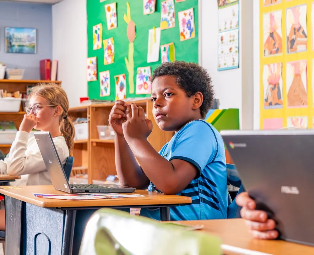 Students sitting at desks in a classroom, with colorful artwork on the walls. One boy wearing a blue shirt is looking attentively, while a girl with glasses and a ponytail is sitting nearby, focused.