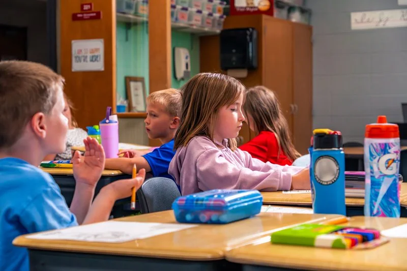 Young students sitting at desks in a classroom, engaged in learning, with school supplies on their desks.