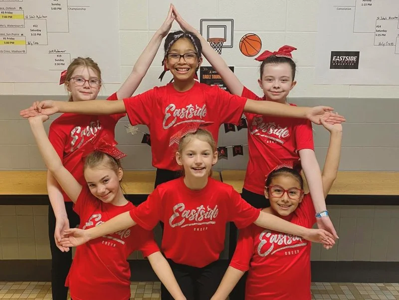 Five young girls in red Eastside Cheer uniforms posing in a gym, smiling and forming a star shape with their arms in front of a wall with sports-themed posters and a basketball hoop illustration.