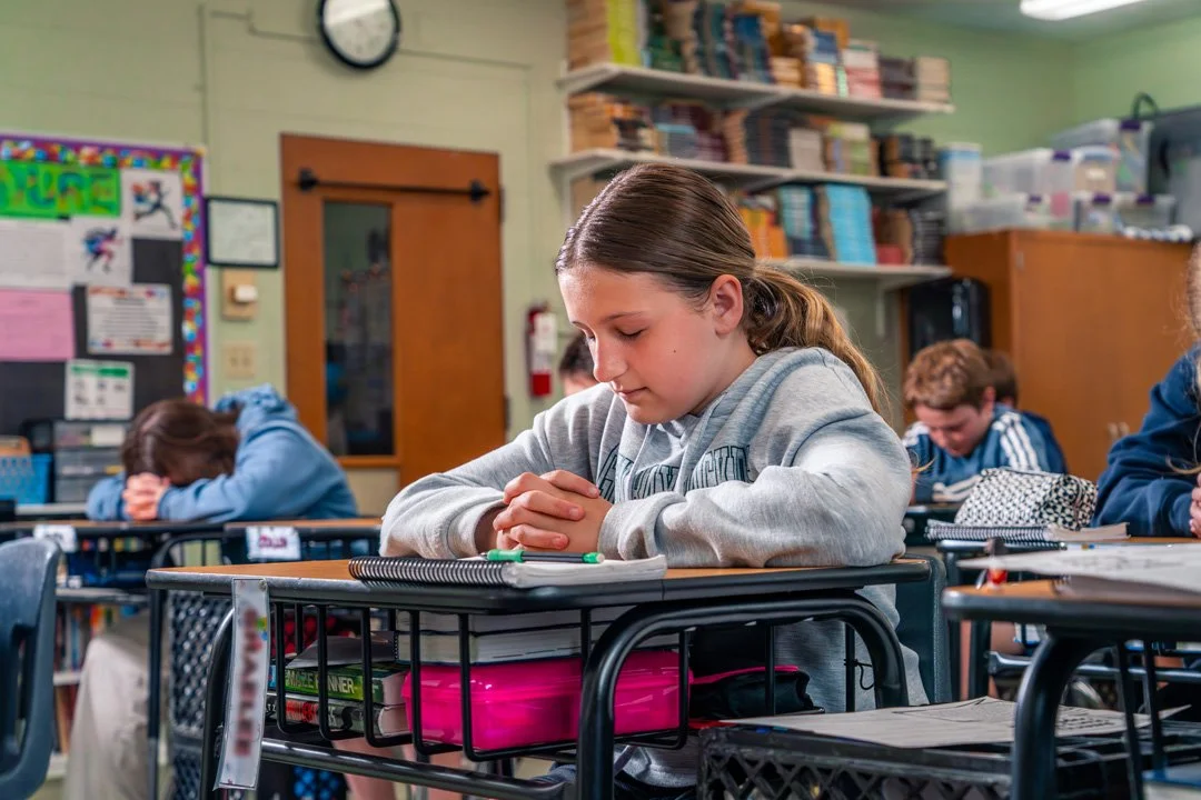 A young girl with brown hair in a ponytail in a classroom, sitting at a desk with hands clasped and eyes closed, next to a pink pencil case and notebooks.