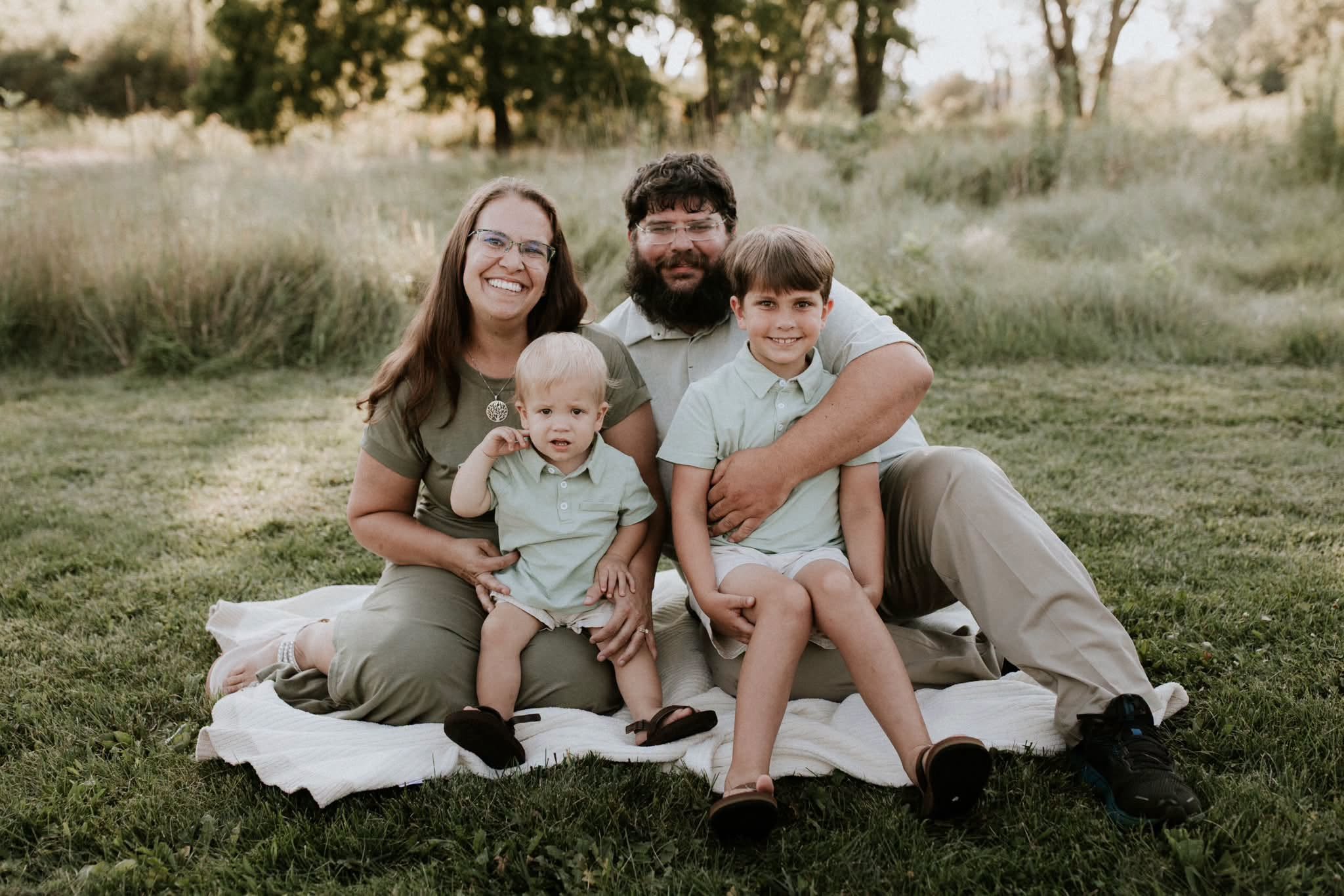 A family of five sitting on a blanket in a park with trees and grass in the background.