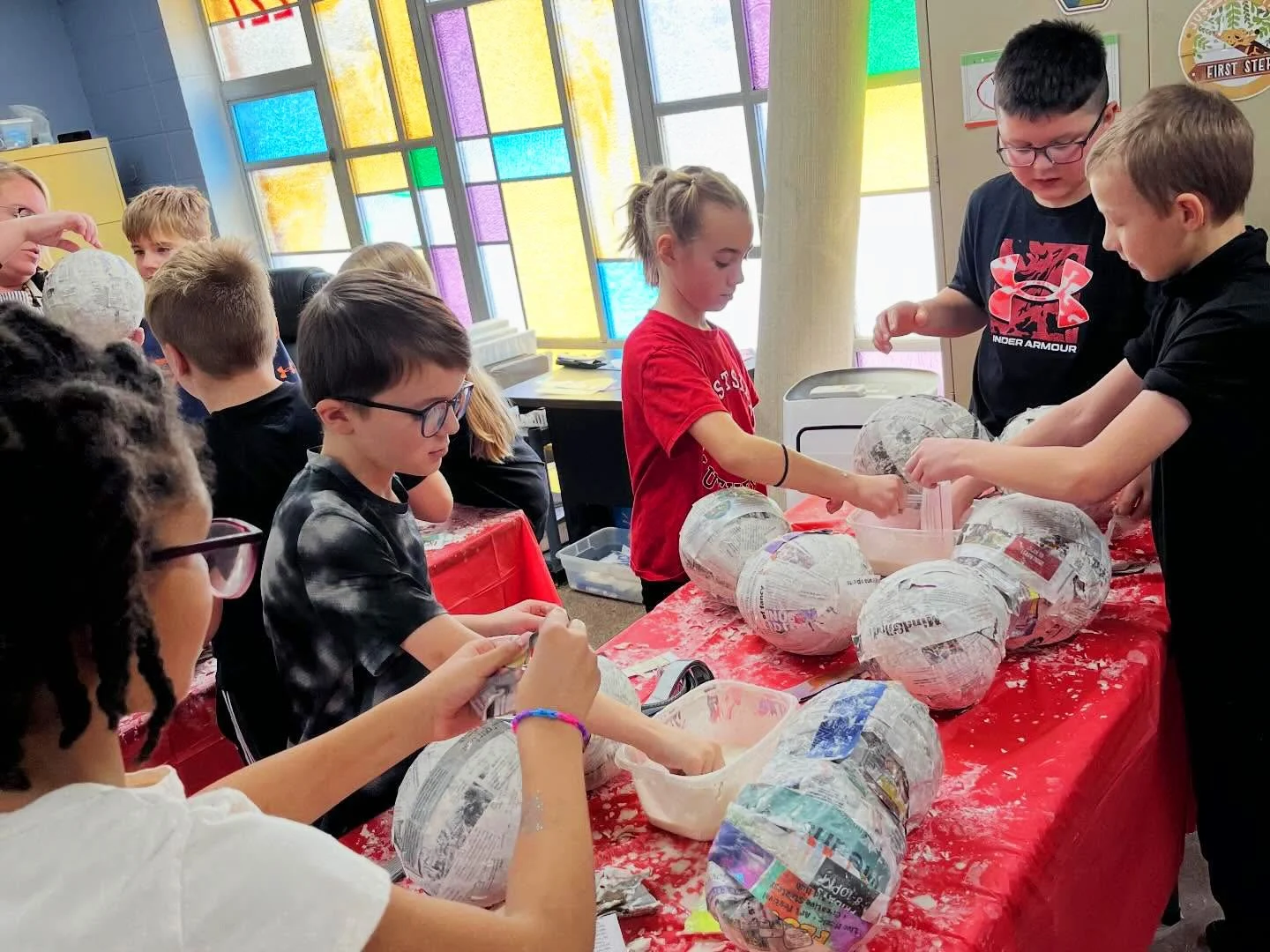 Children creating papier-mâché globes in a classroom with colorful stained glass windows.