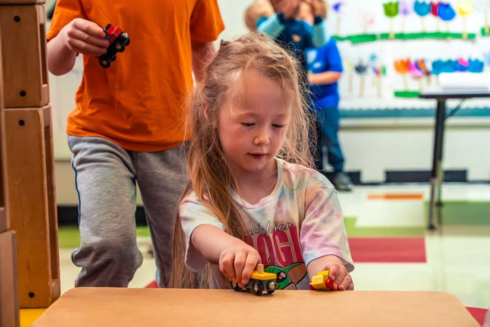 A young girl playing with small toy vehicles at a table in a classroom or play area with colorful decorations and two other children in the background.