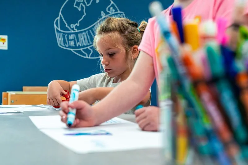 A young girl coloring at a table with markers, with a pile of colorful markers in the foreground and a dark blue wall in the background.