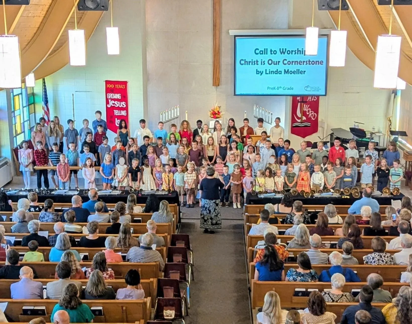 A large group of children on stage in a church, singing or performing, in front of a seated congregation. A woman, likely the choir director or teacher, is conducting them. The church has a high ceiling with hanging lights and a large screen displaying the title and details of the performance. There are banners on the walls and an American flag in the background.