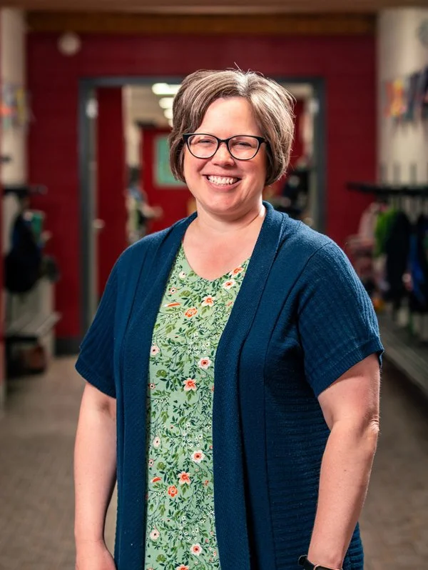 A smiling woman with gray hair, glasses, wearing a floral green dress and a blue cardigan, standing inside a retail store.
