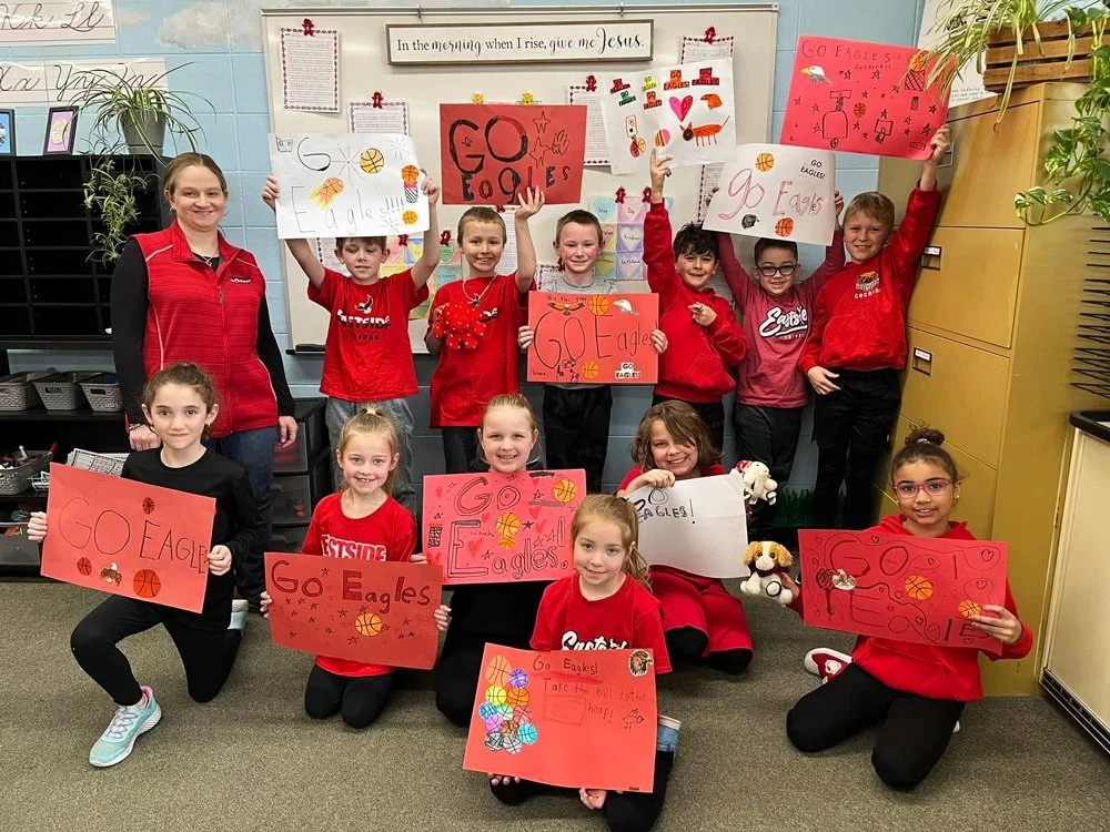 Group of children and a teacher in a classroom holding handmade signs and posters supporting the Eagles school team with football motifs and uplifting messages.