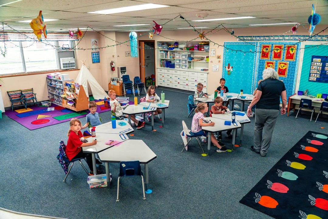 A colorful kindergarten classroom with children sitting at desks, a teacher walking through, and decorations hanging from the ceiling.