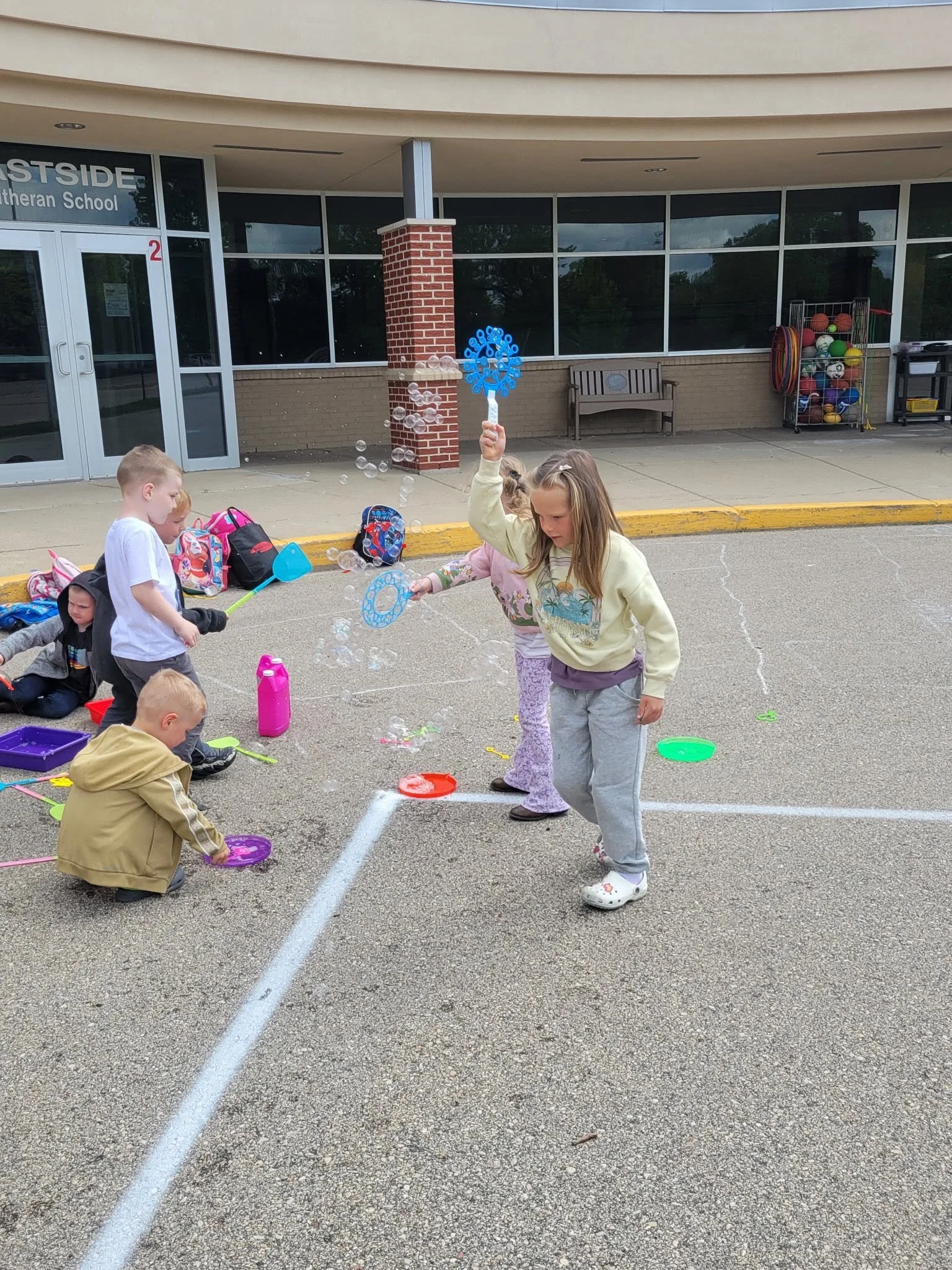 Children playing with bubbles outside of a school building, which has large windows, a brick column, and a sign that reads 'EASTSIDE Lutheran School' in the background.