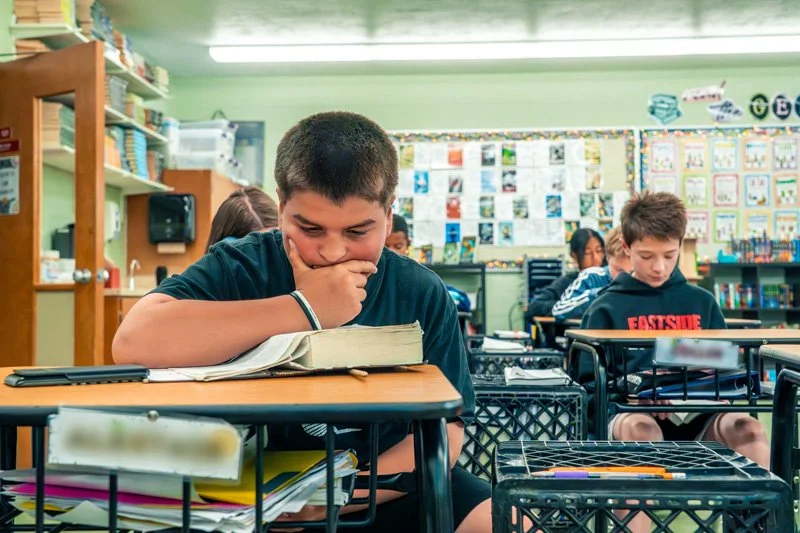 A boy sitting at a desk in an Eastside Lutheran school classroom, reading a book and resting his chin on his hand, with other students and classroom decorations in the background.