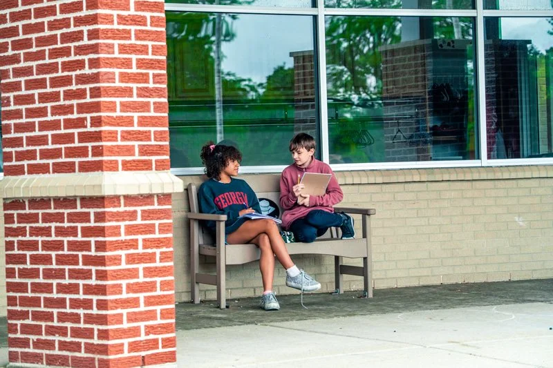 Two students sit on a bench outside a brick building with large windows. One girl wears a navy Georgia sweatshirt and shorts, holding a notebook, while a boy wears a maroon hoodie, holding a cardboard with notes.