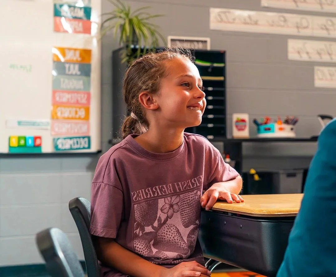 A young girl with braided hair, sitting at a desk in a classroom, smiling and talking to a person out of frame.