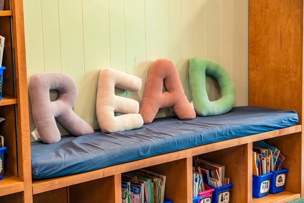Colorful plush pillows spelling out "READ" on a blue cushioned bench in a cozy reading nook, with books stored beneath.