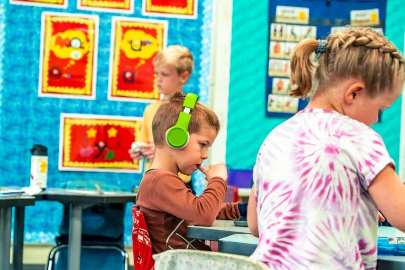 Children sitting at desks in a classroom, with one boy wearing large green headphones, and colorful artwork on the bulletin boards in the background.