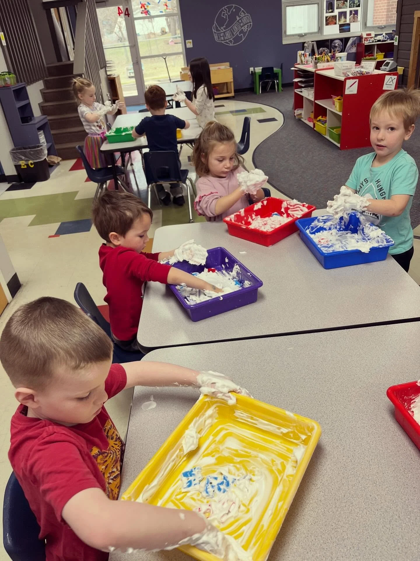 Children in a classroom playing with and exploring white foam or shaving cream on trays and tables.