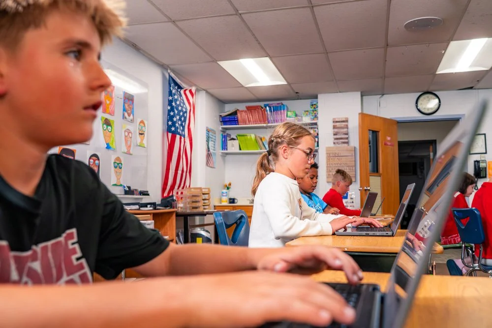 Students working on laptops in a classroom with American flag and educational posters on the walls.