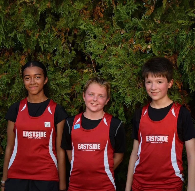 Three young athletes standing outdoors in front of green foliage, wearing red and black sports jerseys that say 'Eastside Lutheran'.
