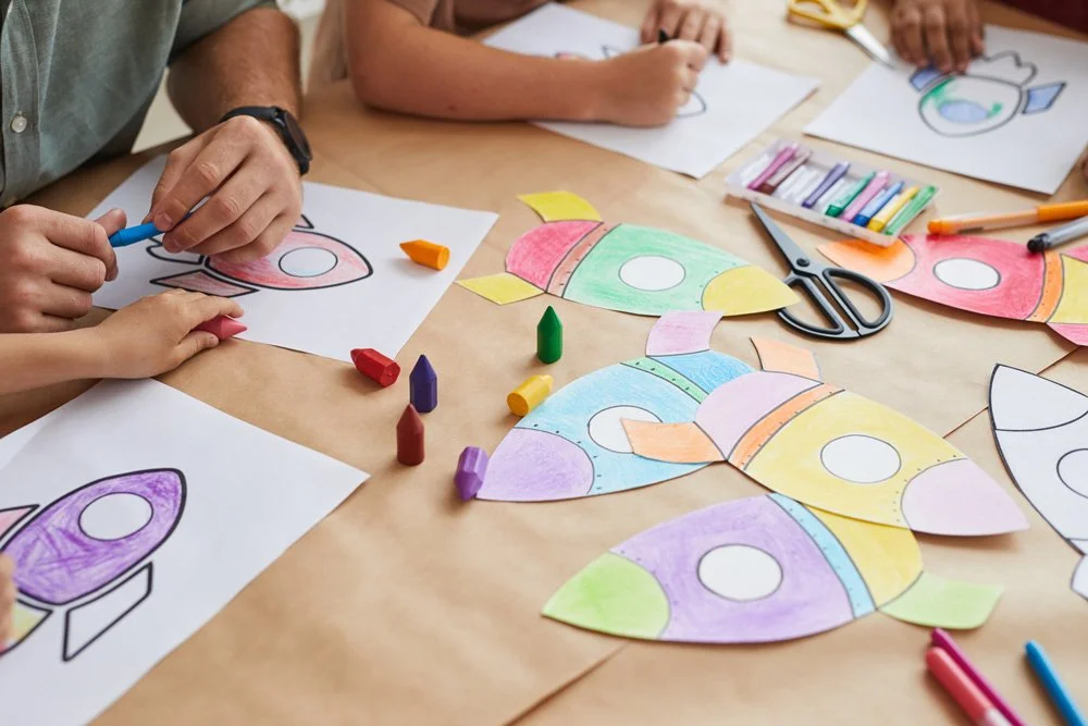 Children and adults coloring firework-shaped paper crafts with crayons and markers on a table.